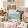 A styled baby gift setup featuring a white and mint green 'Mason Bottle Healthy Baby Starter Set' box in the center, surrounded by wrapped presents, balloons, flowers, candles, and a wooden sign that reads 'Welcome to the World, LITTLE ONE'.